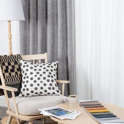 Cru grå mörkläggningsgardin i stilren miljö med ljus träfåtölj, prickig kudde, pläd, kaffekopp och tygprover på runt soffbord.