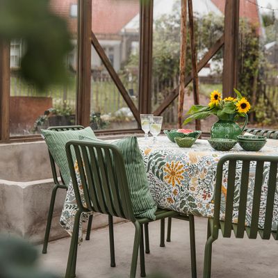 Myllra gul bordsduk på trädgårdsbord i växthusmiljö med gröna stolar och vas med solrosor