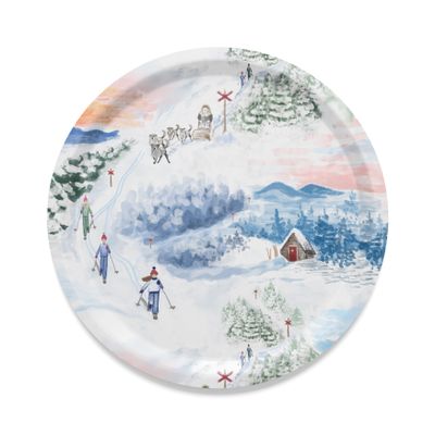 Gnistrande naturvit rund bricka Ø38 cm med skidåkare, hundspann, snötäckta granar, fjäll, stuga och röda ledkryss i ett nordiskt vinterlandskap.