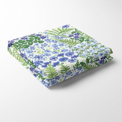 Närbild på den vikta bordsduken Blå Blom i ljusblått med tryckta blå blommor och grönt bladverk, liggandes på vit bakgrund.