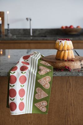 Kökshandduk med äpplen och pepparkakor i rött och grönt, bredvid en sockerkaka dekorerad med lingon.