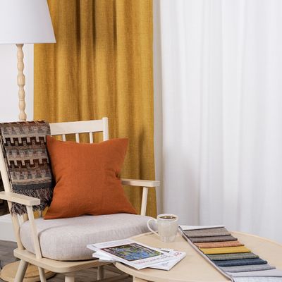 Cru ockra mörkläggningsgardin i dubbel bredd i varm miljö med ljus träfåtölj, rostfärgad kudde, pläd, kaffekopp och tygprover på runt soffbord.
