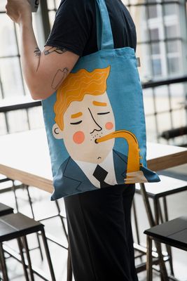 Person bär blå tygkasse med illustrerad saxofonspelare över axeln i ljus inomhusmiljö