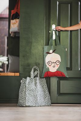 Grön tygkasse med motiv av Bodil som hänger på en dörr, med en mönstrad väska på golvet bredvid