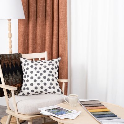 Cru rost mörkläggningsgardin i dubbel bredd i varm vardagsrumsmiljö med ljus träfåtölj, prickig kudde, pläd, kaffekopp och färgprover på soffbord.