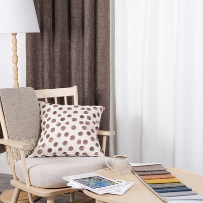 Cru brun mörkläggningsgardin i miljö med ljus träfåtölj, prickig kudde, pläd, kaffekopp och tygprover på runt soffbord.