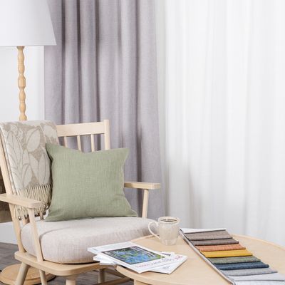 Cru ljusgrå mörkläggningsgardin i stilren miljö med ljus träfåtölj, grön kudde, pläd, kaffekopp och tygprover på runt soffbord.
