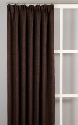 Helkroppsbild av gardinen Milo mörkbrun upphängd vid ett fönster mot en grå vägg med vit spröjs.