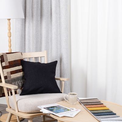 Cru ljusgråmelerad mörkläggningsgardin i dubbel bredd i stilren miljö med ljus träfåtölj, mörkblå kudde, pläd, kaffekopp och tygprover på runt soffbord.