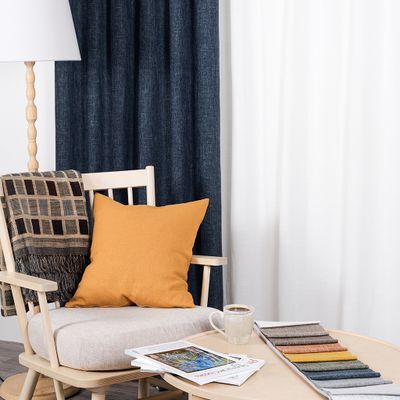 Cru mörkblå mörkläggningsgardin i miljö med ljus träfåtölj, kudde i ockragult, pläd, kaffekopp och tygprover på runt soffbord.