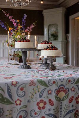 Närbild på Torplycka beige bordsduk med färgstarka blommor och slingrande blad, dukad med tårtor, jordgubbar och ljusstakar.