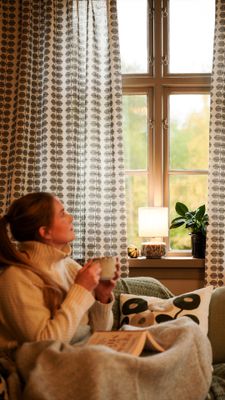 Berså grå gardiner med stiliserat bladmönster på ljus botten vid ett spröjsat fönster. Sofia sitter i en grön fåtölj med en kopp i händerna och en uppslagen bok i knät. På fönsterbrädan står en tänd lampa och en grön krukväxt.