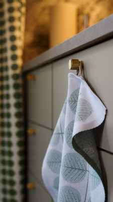 Berså kökshandduk i vitt med gröna blad, hängande i mjuka veck från en rund mässingsfärgad knopp. Närbilden visar handdukens vävda struktur och bladmönster, med köksskåp och matchande gardin från samma serie i bakgrunden.