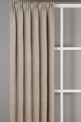 Owen Beige gardin från Fondaco upphängd framför en grå vägg med vit spröjsad inredningsdetalj i bakgrunden. Gardinen faller i långa, mjuka veck och den ribbade strukturen syns över hela tyget.