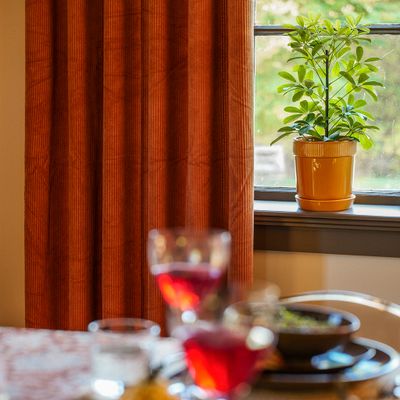Owen rost manchestergardin från Fondaco i varm hemmiljö, hängande bredvid ett fönster med en grön krukväxt på fönsterbrädan och ett dukat bord framför.