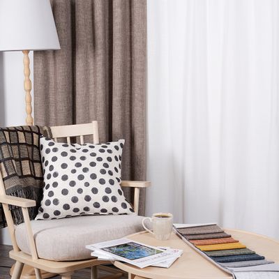 Cru mullvad mörkläggningsgardin i hemtrevlig miljö med ljus träfåtölj, prickig kudde, pläd, kaffekopp och tygprover på runt soffbord.