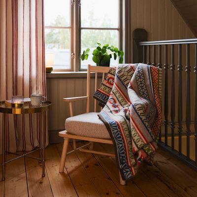 Kerstin Landström Design ullfilt Skaraborg draperad över en träfåtölj i en ombonad miljö med trägolv. I bakgrunden syns den randiga gardinen Hedvig offwhite, en grön växt och en liten bordslampa.