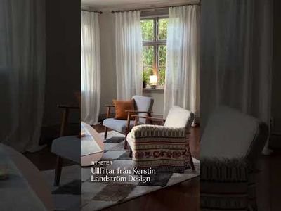 Miljöbild med en mönstrad ullfilt från Kerstin Landström Design draperad över en fåtölj, med ytterligare en fåtölj, orange kudde, ljusa gardiner och ett fönster i bakgrunden.