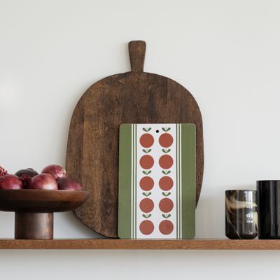 Uppläggningsbrädan Frukt och kaka i rödgrönt står lutad mot en mörk träbräda på en hylla, omgiven av rödlök, glas och köksdetaljer i ett modernt hem.