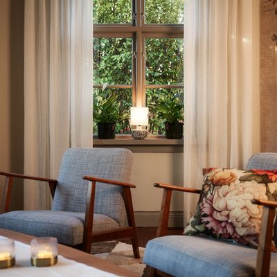 Brooke offwhite gardiner från Jakobsdals inramar ett stort fönster i en ombonad vardagsrumsmiljö med fåtöljer och gröna växter.