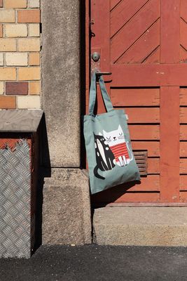 Tygkasse med kattmotiv hänger på ett dörrhandtag vid en röd trädörr utomhus