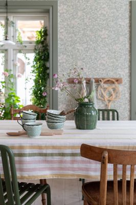 Alba randig bordsduk på matbord i ljus matsal med gröna koppar, skålar och vas med blommor.