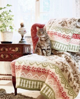 Torpet ullfilt draperad över röd fåtölj i lantligt vardagsrum med spetsgardiner och katt sittande på filten.
