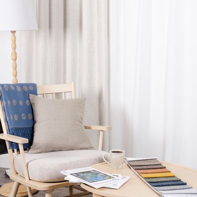 Cru offwhite mörkläggningsgardin i lugn vardagsrumsmiljö med ljus träfåtölj, beige kudde, blå pläd, kaffekopp och färgprover på soffbord.