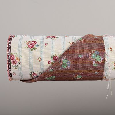 Närbild på Romie beige jacquardvävt möbeltyg på rulle där både framsidans romantiska rosmönster och den invävda baksidan syns.