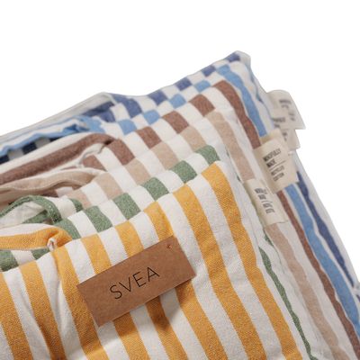 Närbild av Svea sittdynor i flera färger med randigt mönster i vitt och gula, gröna, beige, bruna och blå nyanser. Fokus ligger på den gula sittdynan med etikett och synliga detaljer i tyget och handtaget.