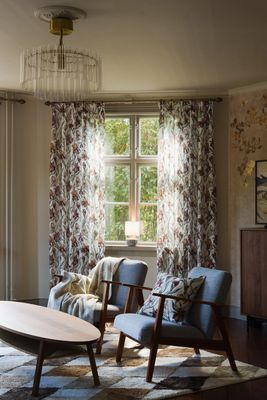 Ljust vardagsrum med de mönstrade gardinerna Iris i färgen offwhite, matchande kuddfodral Iris och en ljus pläd. Gardinerna ramar in fönstret och skapar en harmonisk helhet tillsammans med rummets varma trädetaljer och dämpade färgskala.
