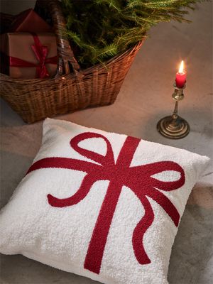 Paket kuddfodral i vit teddy med mörkröd rosett placerat på golv intill julgran och ljusstake