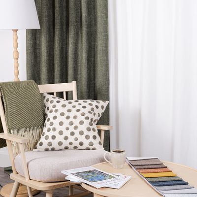 Cru grön mörkläggningsgardin i dubbel bredd i harmonisk miljö med ljus träfåtölj, prickig kudde, grön pläd, kaffekopp och tygprover på runt soffbord.