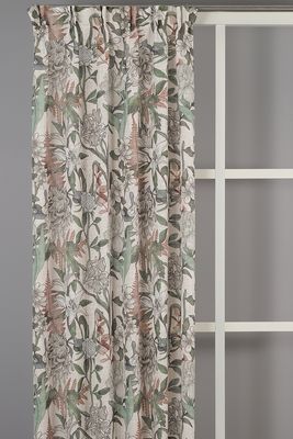 Malou Beige gardin från Redlunds upphängd framför en grå vägg. Det storskaliga mönstret med blommor, blad och växtmotiv följer gardinens långa veck och visar hur motivet upplevs över en större yta.