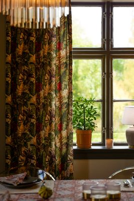 Blommiga Yucatan-gardiner med stora gröna blad och blommor i rött, gult och cremevitt vid ett spröjsat fönster. På fönsterbrädan står en grön krukväxt, ett ljus och en bordslampa. I förgrunden syns ett dukat